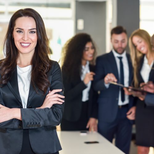 Businesswoman leader looking at camera in modern office with multi-ethnic businesspeople working at the background. Teamwork concept. Caucasian woman.